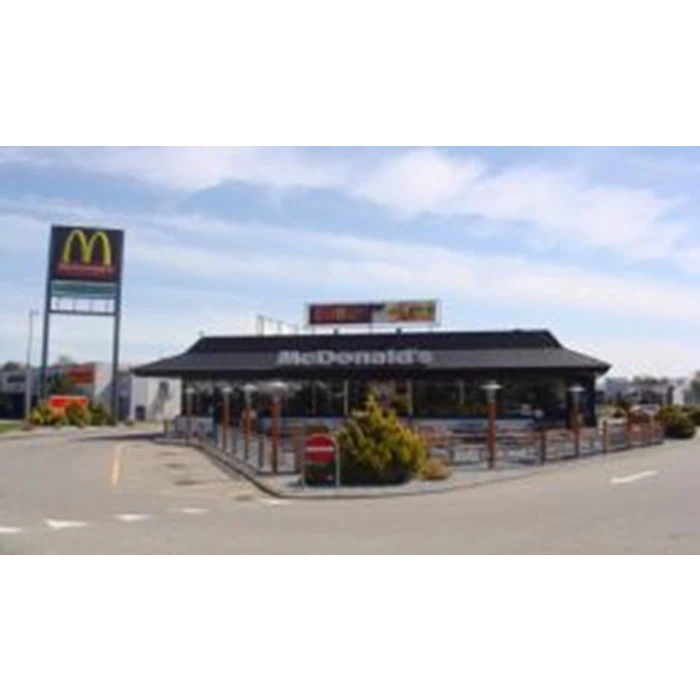 En McDonalds-restaurant med udendørs siddepladser, en drive-thru-bane og et højt skilt med McDonalds-logoet nær parkeringspladsen.